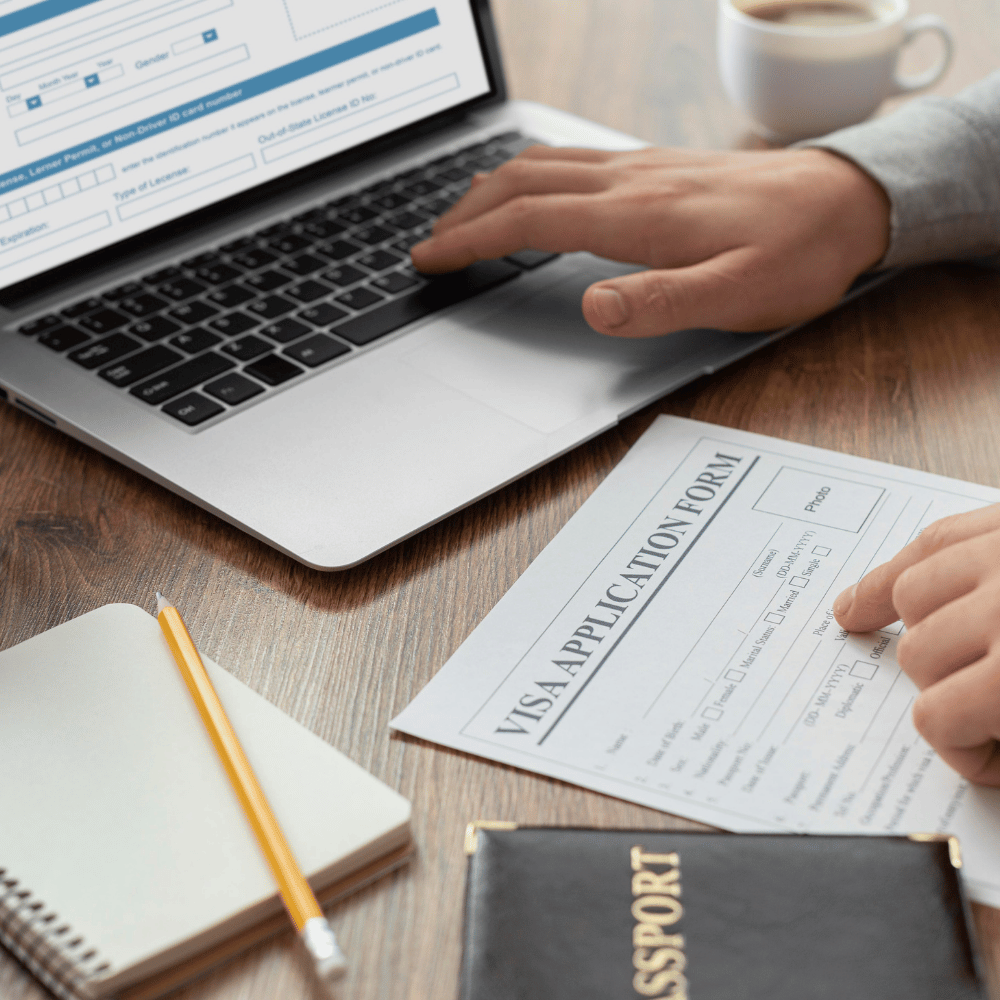 Hands filling out a visa application form next to a laptop and passport on a wooden desk, representing visa consulting services in Greater Noida